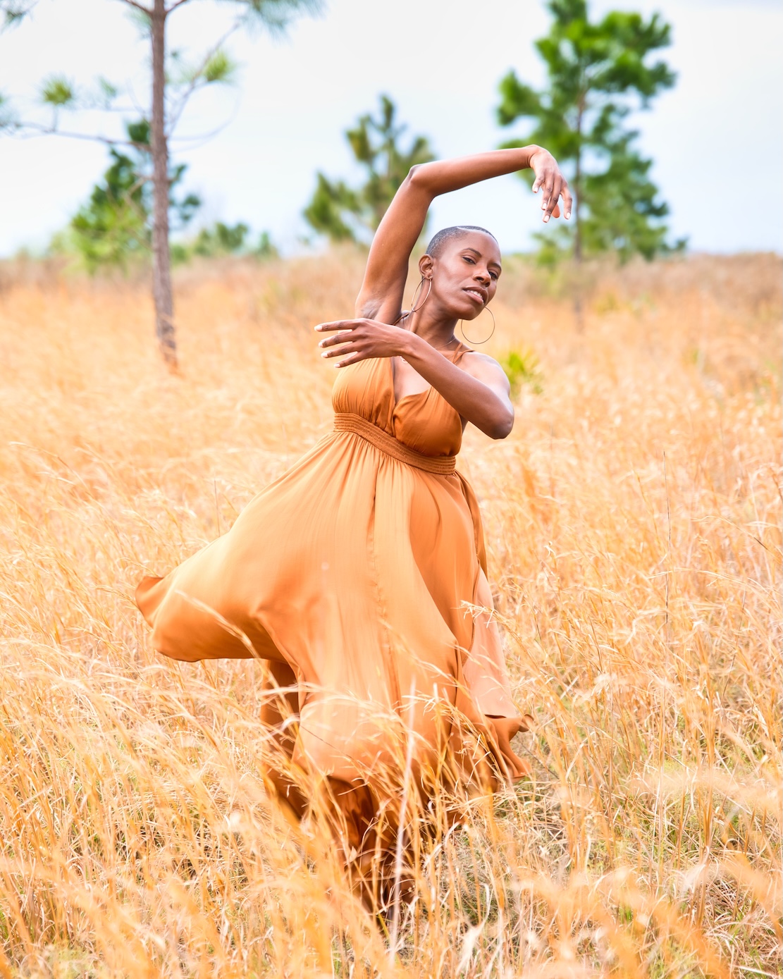 an African woman in an orange dress posed with arms raised and one leg up, in the middle of a field with trees in the back of her