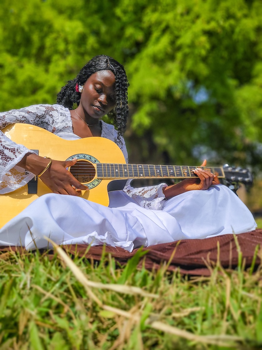 an African woman in a white dress is sitting cross-legged on a brown cloth, strumming a guitar