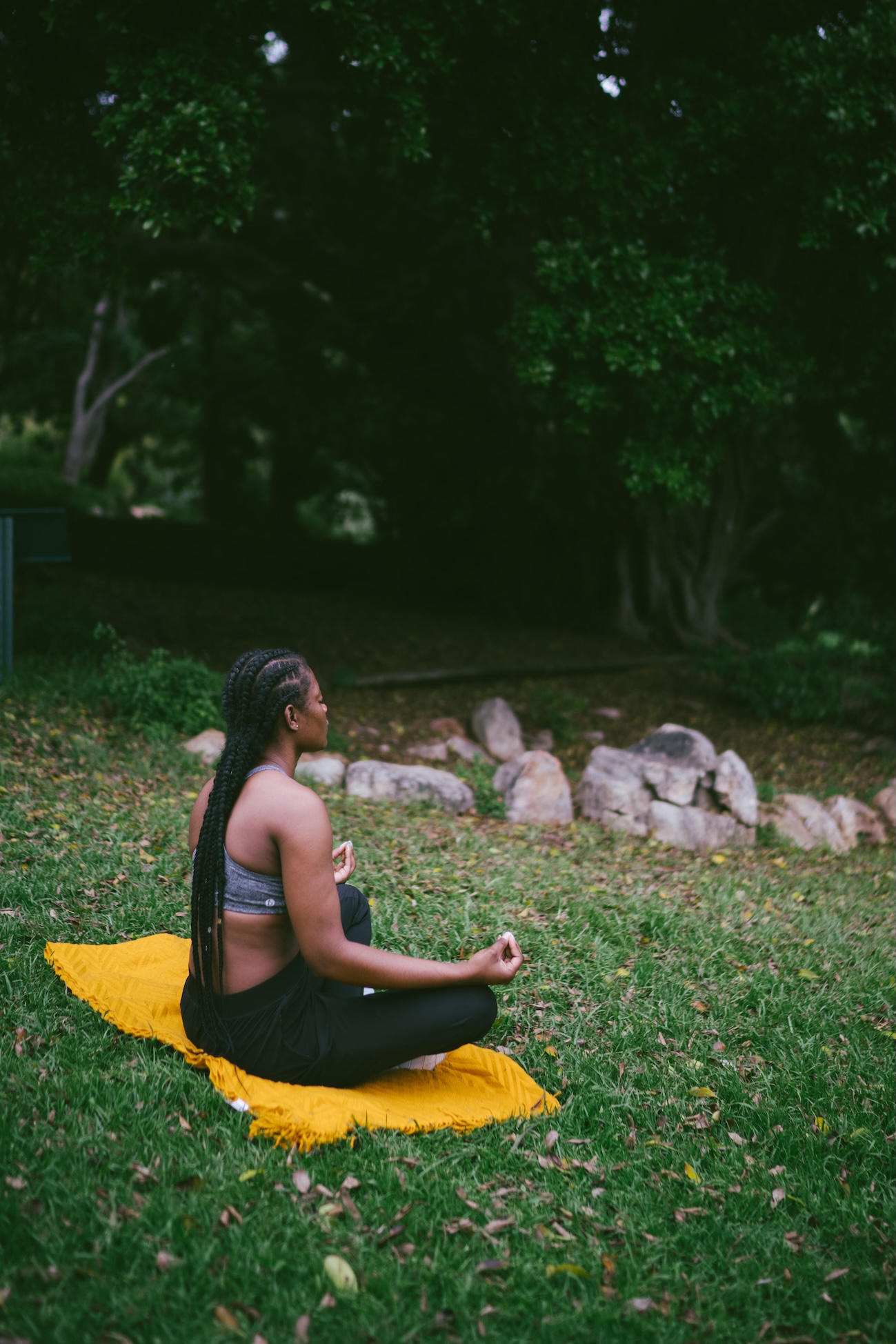 an African woman sitting cross-legged on a mustard yellow cloth, on a grassy field facing away from the camera